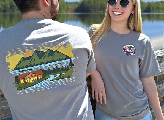 Two people wearing gray t-shirts with scenic designs, one featuring a lake and cabin, the other with a logo.