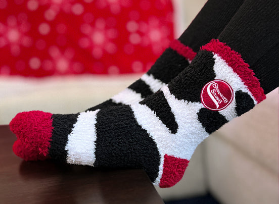 A photo of feet wearing cozy black, white, and red fuzzy socks with a cow-print design. The socks feature a red circle with the Stewart's Shops logo on the side. The feet are propped up on a table, with a red blanket featuring a snowflake pattern in the background, creating a warm and relaxed atmosphere.