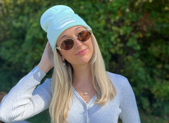 Woman wearing a light blue beanie and sunglasses with a blurred green background