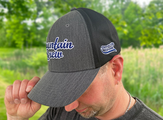 A person wearing a dark gray baseball cap with "Mountain Brew" embroidered in white and blue script on the front. The person is outdoors in a grassy field with trees in the background and is tipping the brim of the cap downward with one hand. The Stewart's logo is visible on one side of the black panels.