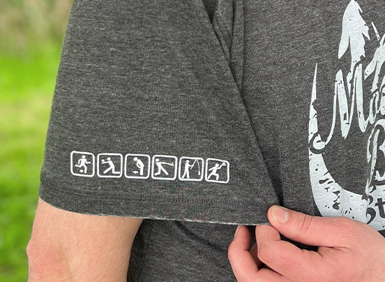 Close-up of a person wearing a dark gray t-shirt with the left sleeve pulled slightly forward to show a row of six small white icons. The icons depict outdoor activities: running, biking, hiking, kayaking, skiing, and snowboarding. Part of the "Mountain Brew Stewart’s" graphic on the shirt front is also visible.