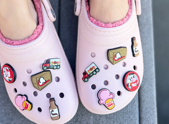 Image of pink clogs adorned with Stewart's Shops-themed shoe charms. The charms include designs such as a milk carton, a pink ice cream cone with the Stewart's Shops logo, a delivery truck, a chocolate milk bottle, and a circular charm featuring a cartoon cow. The shoes also feature pink fuzzy liners visible at the edges.