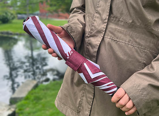 A person in a tan jacket holds a closed maroon and white Stewart’s Shops umbrella while standing near a pond on a cloudy day. The umbrella is neatly wrapped and secured with a matching strap.
