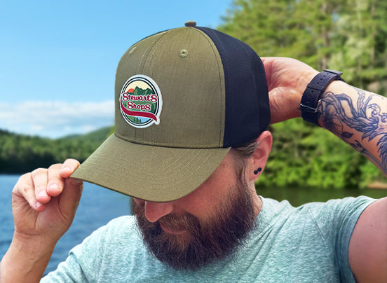 Bearded man wearing an olive green and black Stewart’s Shops trucker-style hat while standing near a lake with a forested shoreline in the background. The hat features a round patch with mountains, trees, a sunset, and the Stewart’s Shops logo. The man is looking down slightly and adjusting the brim of the hat with both hands.