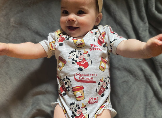 Smiling baby wearing a gray Stewart’s Shops onesie with an all-over print design featuring cartoon cows, ice cream pints, and the phrases “Pint Sized” and “Stewart’s Shops.” The baby is lying on a soft gray blanket with arms outstretched.