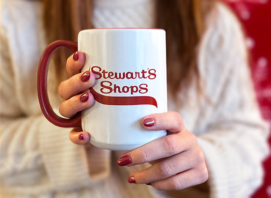 Close-up of a person holding a white ceramic mug with the Stewart's Shops logo, featuring a red handle and red painted nails, set against a cozy background