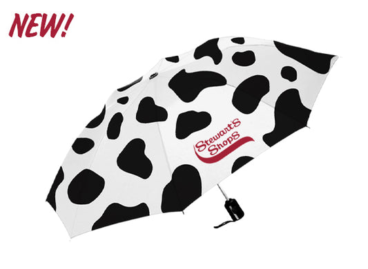 White compact umbrella with a black-and-white cow print pattern and the red Stewart's Shops logo printed on one panel. The umbrella is shown open against a light gray background, with a "NEW!" callout in red script in the upper-left corner. The folded black handle and wrist strap are visible beneath the canopy.