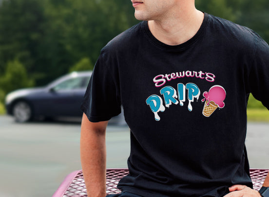 A man wearing a tshirt that says Stewart's Drip with an ice cream cone. He is sitting on a picnic table.