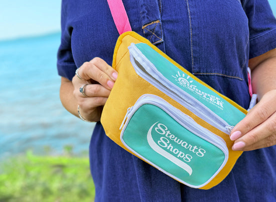 Person holding a yellow and teal waist bag with 'Stewart's Shops' branding by a body of water.