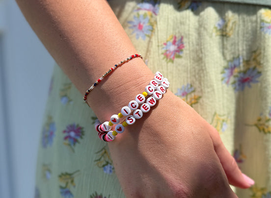 Close-up of a wrist wearing two colorful bracelets with letters, against a floral-patterned background.