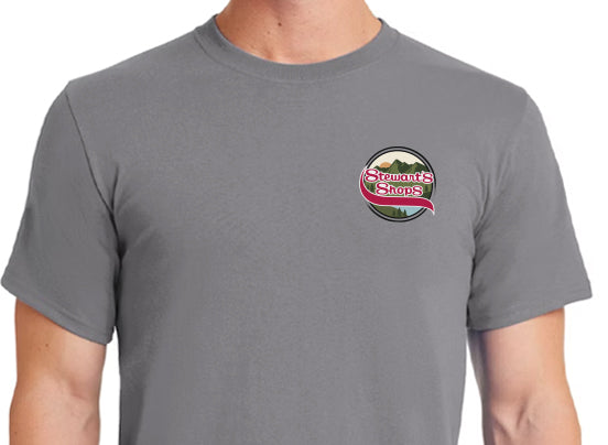Gray short-sleeve t-shirt worn by a person. The shirt features a small circular Stewart’s Shops logo on the left chest, with a red ribbon-style banner and mountain background inside the circle.