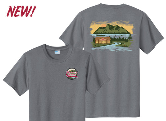 Gray short-sleeve t-shirt with front and back designs. The front features a small circular Stewart’s Shops logo on the left chest. The back showcases a colorful scenic illustration of a Stewart’s Shops building by a lake, surrounded by trees with mountains and a sunset sky in the background. The word “NEW!” appears in red text at the top left corner.