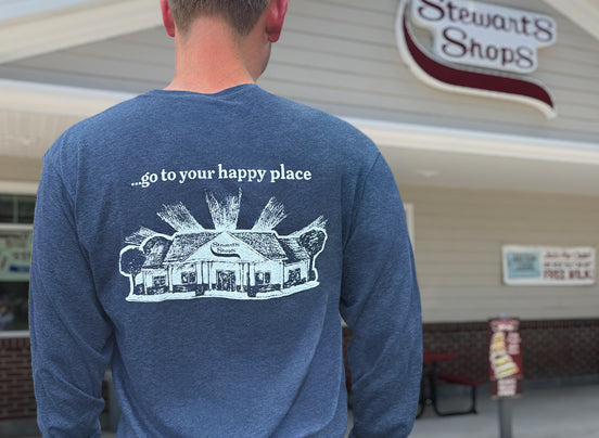Person wearing a blue Stewart’s Shops long-sleeve shirt featuring a Stewart’s shop illustration and the phrase “…go to your happy place” on the back, standing outside a Stewart’s Shops location.