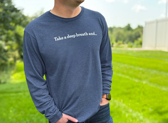 Person wearing a navy blue long-sleeve shirt with white text across the chest reading “Take a deep breath and…” while standing outdoors.
