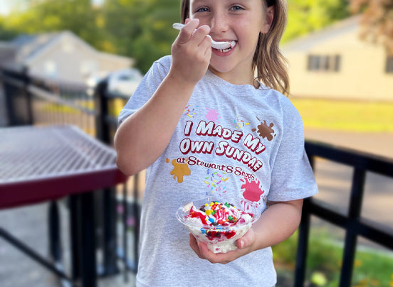 Child eating ice cream with a 'I Made My Own Sundae' shirt on a sunny day.