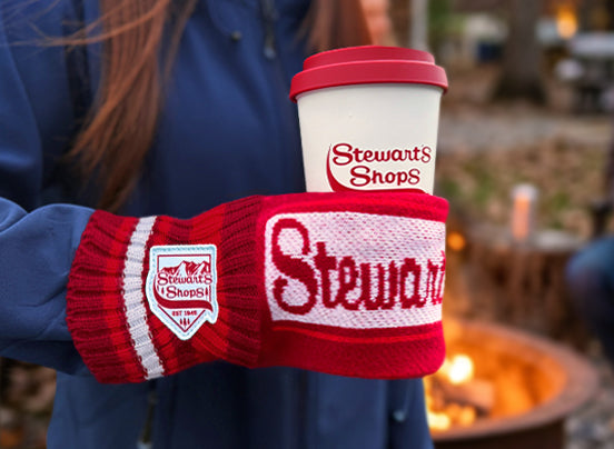 One person wearing a knit mitten can holder holding a coffee cup. There is a Stewart's Shop logo on the mitten and the words Stewart's Shops knit into the front. 