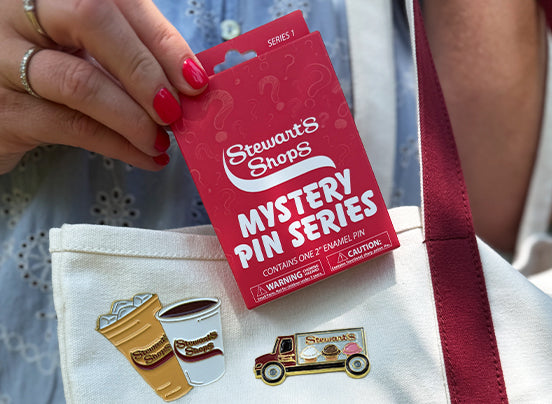 Hand holding a Stewart's Shops Mystery Pin Series package above a tote bag displaying collectible enamel pins of Stewart's coffee cups and a Stewart's delivery truck.