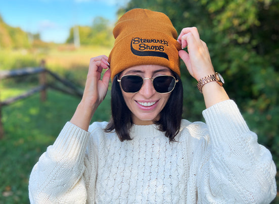 Person wearing a brown beanie with 'Stewarts Shops' logo and sunglasses, standing outdoors.