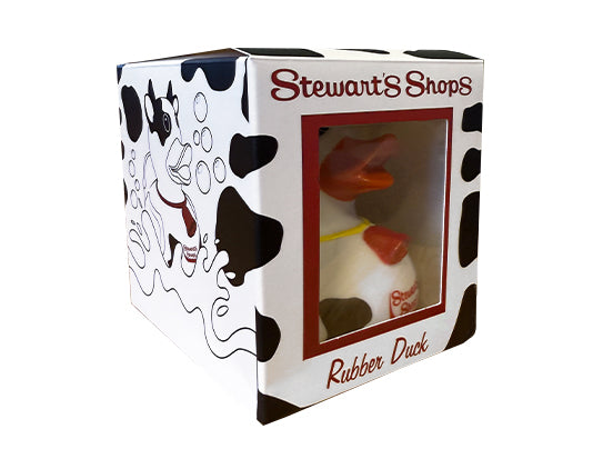 A boxed rubber duck from Stewart’s Shops. The packaging features cow-print designs and an illustration of the duck blowing bubbles. The front of the box has a clear window showing the duck inside, which has a white body with black cow spots, an orange bill, and a red Stewart’s Shops logo. The text “Stewart’s Shops” is printed at the top, and “Rubber Duck” is written at the bottom of the front panel.