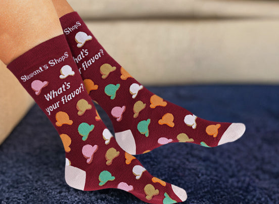 Person wearing Stewart's Shops burgundy crew socks featuring colorful ice cream scoop patterns and the phrase "What's your flavor?" while relaxing indoors.