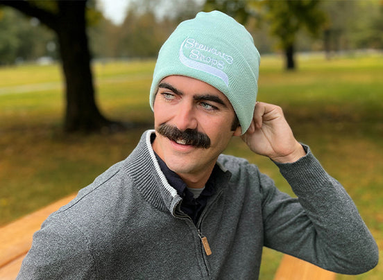 Man wearing a light blue beanie with a logo, sitting outdoors in a park.