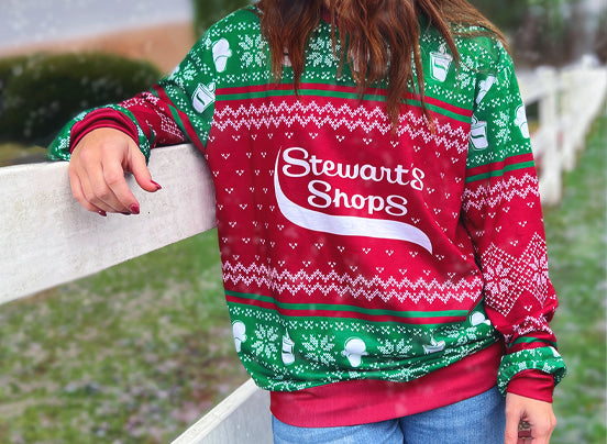 A person leaning on a white wooden fence wearing a festive Stewart's Shops holiday sweater in red, green, and white, featuring a winter-themed pattern with ice cream cones, coffee cups, and snowflakes. 