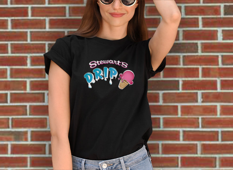 A woman wearing a t-shirt that says Stewart's Drip with an ice cream cone on it. She is wearing sunglasses and is standing in front of a brick wall. 