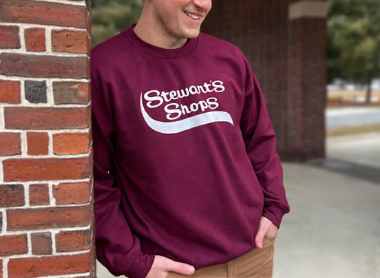 Male wearing burgundy crewneck sweatshirt with Stewart's Shops logo leaning on a brick wall