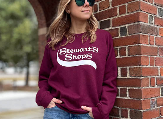 Female wearing burgundy crewneck sweatshirt with Stewart's Shops logo leaning on a brick wall