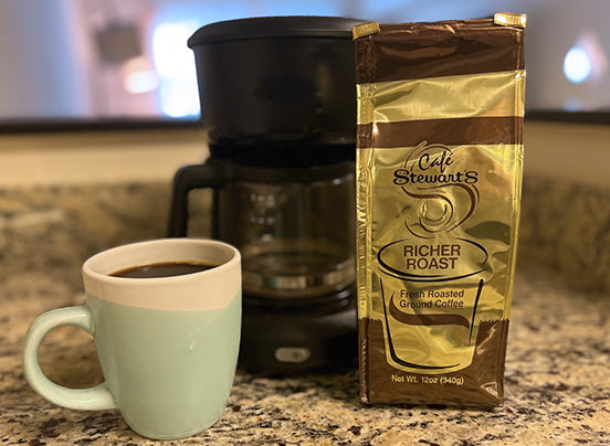 A mug filled with coffee in front of a coffee maker and a 12 oz bag of Stewarts Richer Roast Coffee